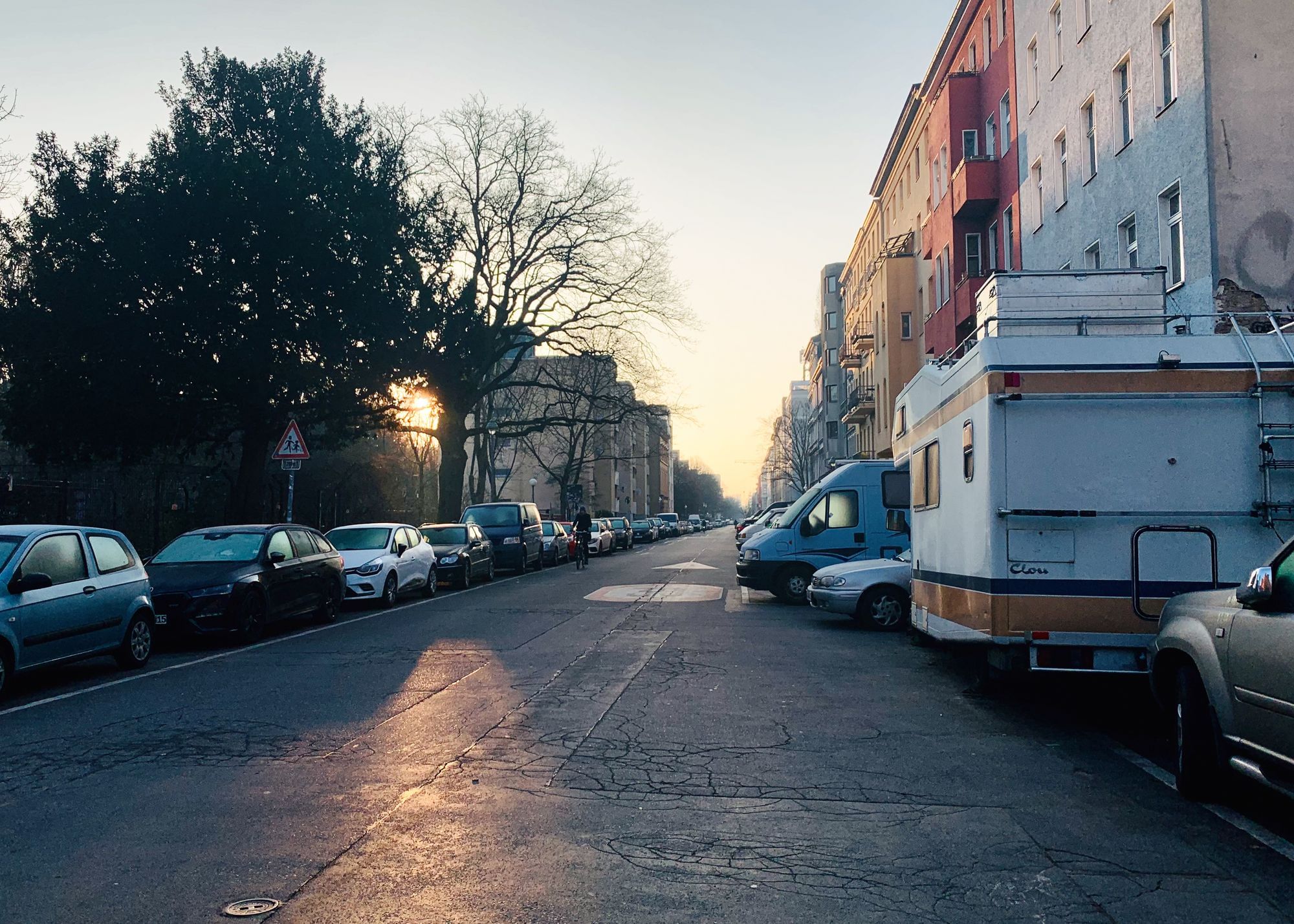 Das Bild zeigt eine ruhige städtische Straße bei Sonnenaufgang. Auf beiden Seiten der Straße parken Autos, darunter ein Wohnmobil. Die Gebäude auf der rechten Seite sind in verschiedenen Farben gestrichen, darunter Rot, Gelb und Weiß. Links säumen Bäume die Straße, und die tief stehende Sonne scheint durch das Laubwerk, was eine warme, goldene Lichtstimmung erzeugt. Ein Straßenschild für einen Fußgängerüberweg ist sichtbar, und die Straße zeigt leichte Risse im Asphalt. Im Hintergrund erstreckt sich die Straße in die Ferne, gesäumt von mehrstöckigen Wohnhäusern.