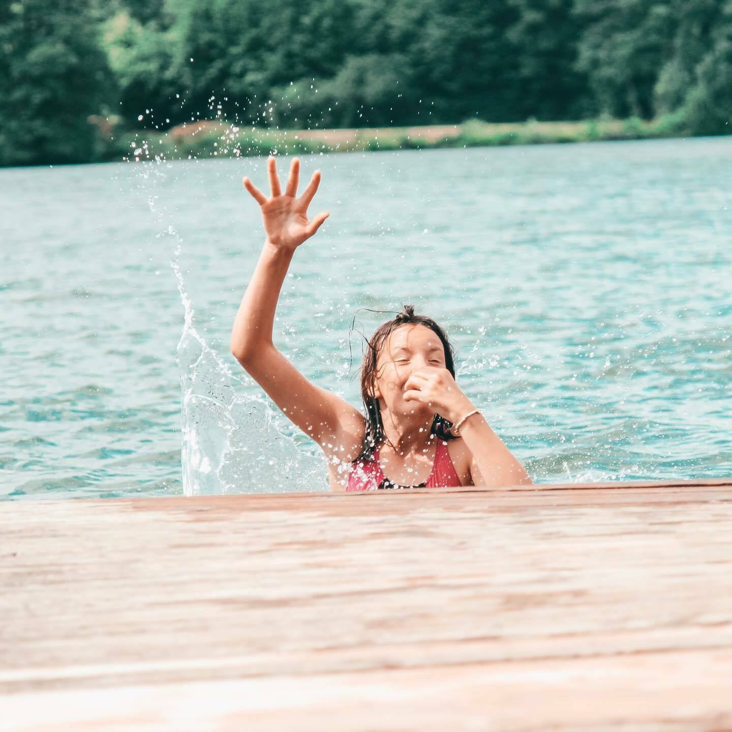 Ein Mädchen ist vom Steg gesprungen in den See. Sie hält sie die Nase zu und reißt den Arm nach oben.