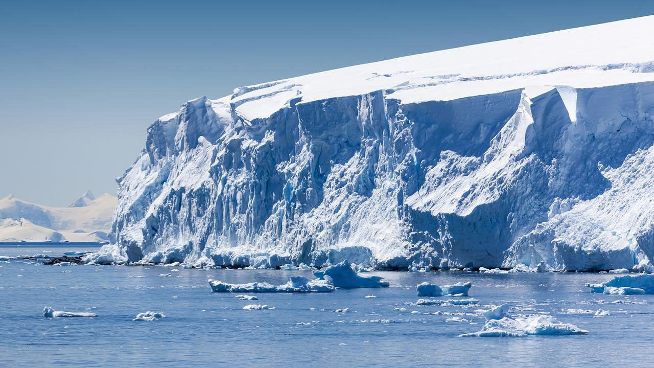 Eine massive Eiswand ragt aus dem Meer, mit scharfkantigen Bruchstellen und Klüften im Gletscher. Im Vordergrund treiben kleinere Eisberge im ruhigen Wasser. Im Hintergrund sind schneebedeckte Berge zu sehen, die den eisigen, klaren Himmel ergänzen. Die Szene vermittelt die Kälte und die majestätische Stille der Polarregion.