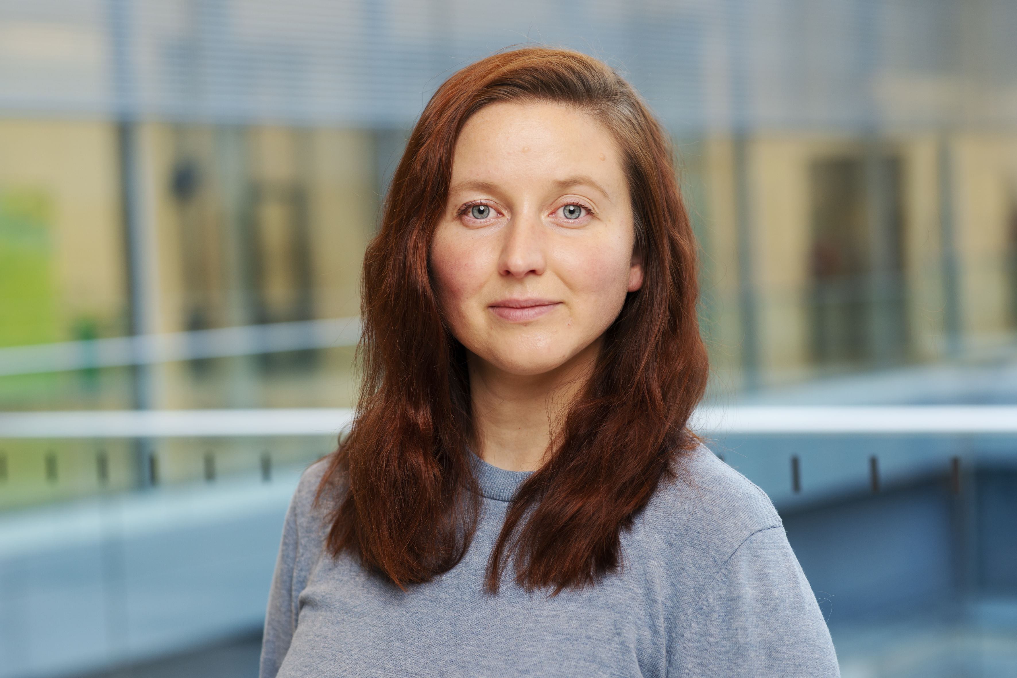 Ein Portrait einer jungen Frau vor einer Glaswand. Sie hat rote Haare und trägt einen grauen Pullover.