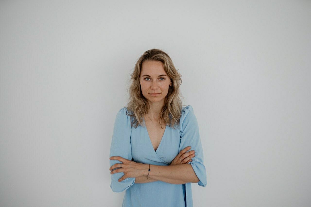 Eine junge Frau steht mir verschränkten Armen vor einer weißen Wand. Sie hat blonde Haare und trägt eine blaue Bluse.