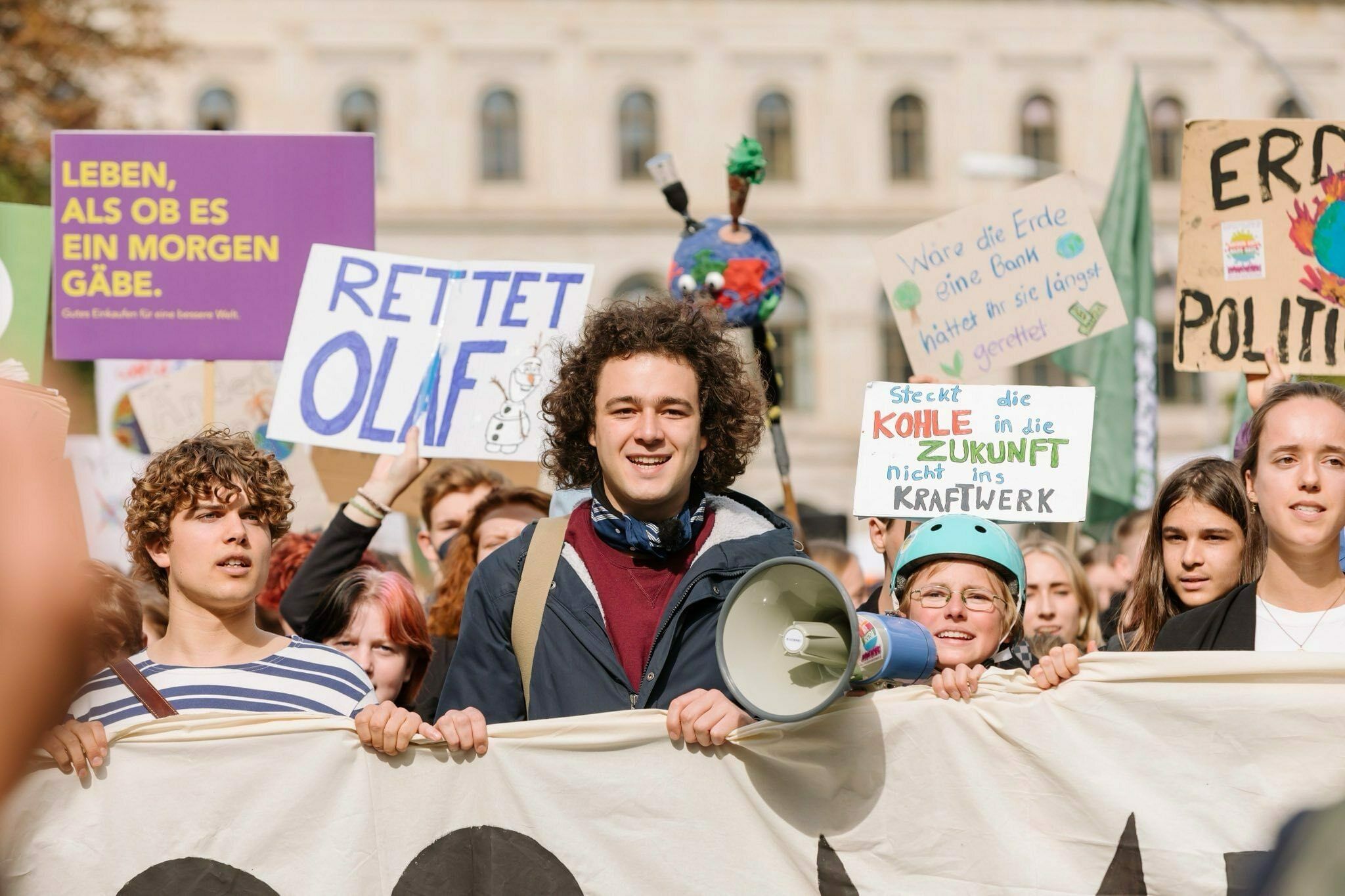 Mehrere Menschen halten auf einer Demonstration ein Transparent in die Höhe. Im Zentrum steht ein junger Mann mit lockigen Haaren. Er trägt einen roten Pulli und eine blaue Jacke.