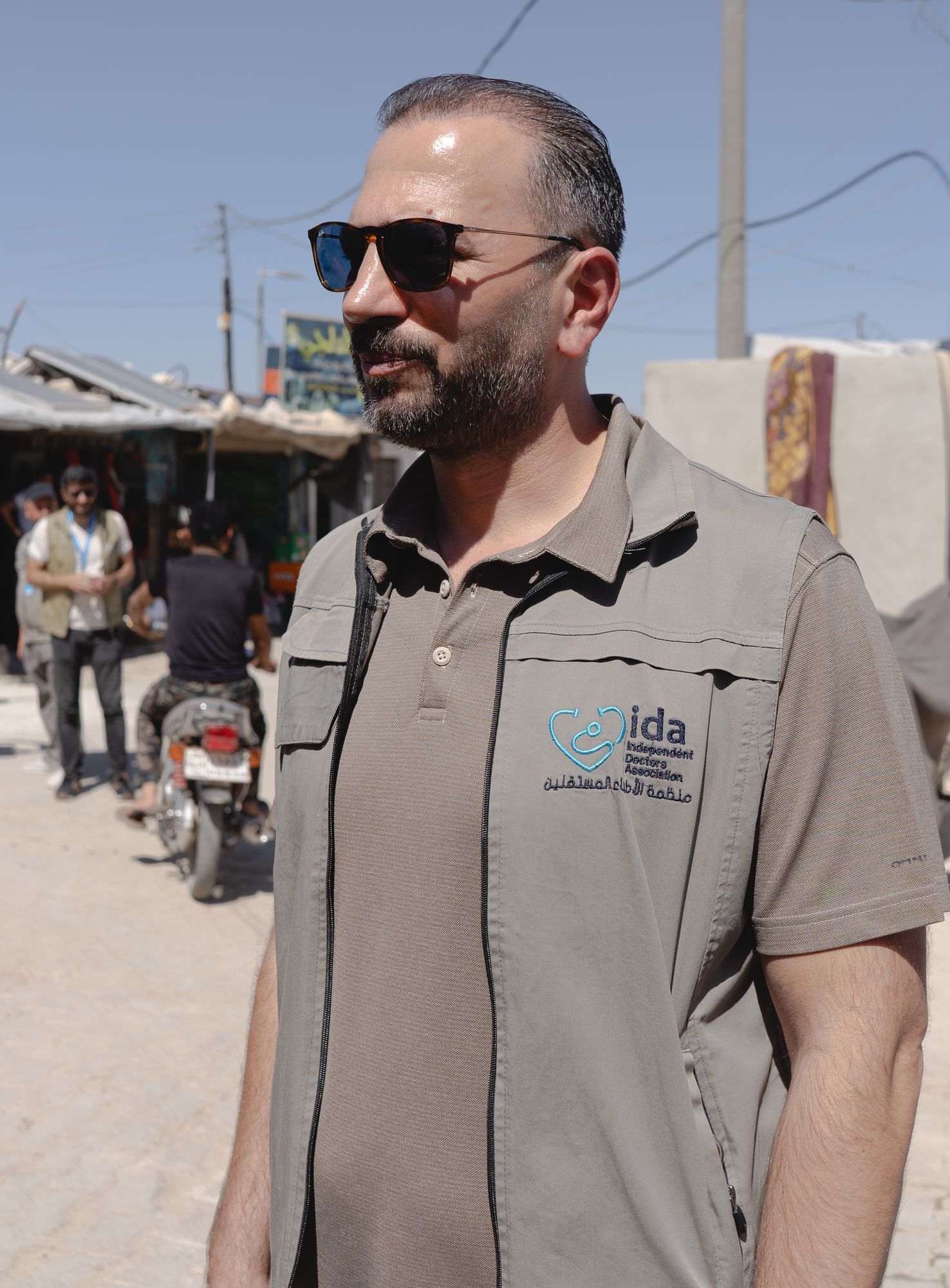 Mahmoud Mustafa trägt ein braunes Poloshirt und eine beigefarbene Weste. Im Hintergrund sieht man einen Menschen auf einem Motorroller und ein Stück einer Mauer. Mahmoud trägt außerdem eine Sonnenbrille.