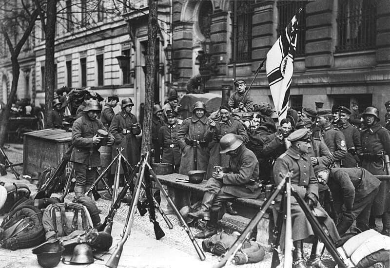 Soldaten der Kapp-Putsch-Truppen rasten auf einer Berliner Straße, 1920. Im Vordergrund aufgestapelte Gewehre und Helme. Die Männer tragen Militäruniformen und eine Reichskriegsflagge ist zu sehen. Historische Fassaden säumen den Hintergrund.
