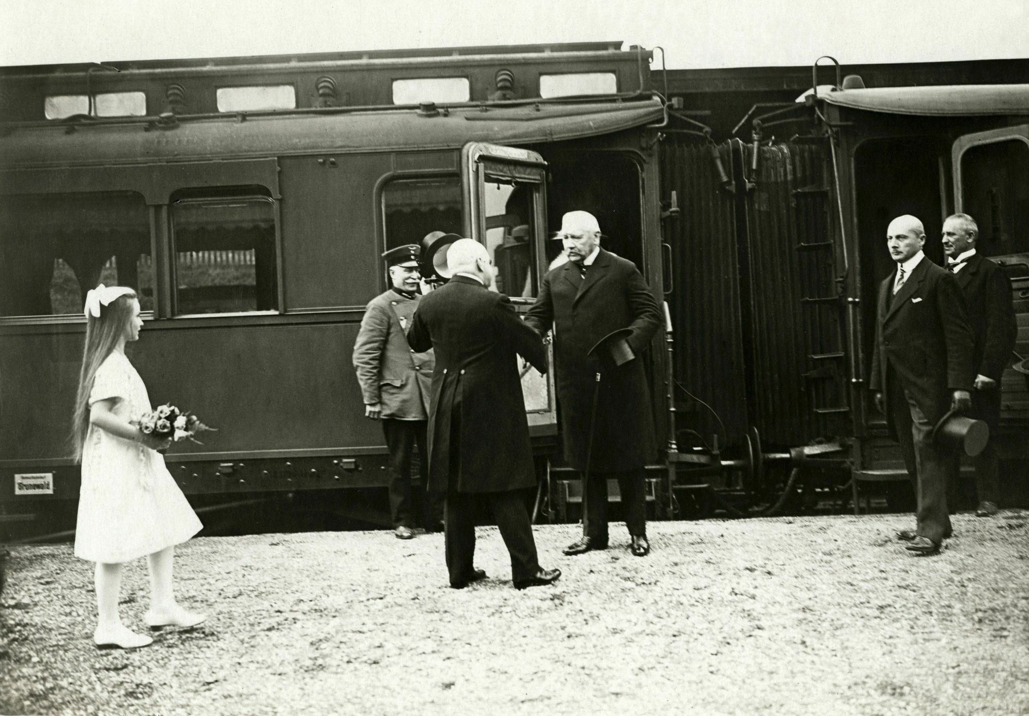 Reichspräsident von Hindenburg wird am Bahnsteig begrüßt. Ein Eisenbahnwaggon bildet den Hintergrund. Ein Mädchen in weißem Kleid mit Blumen steht daneben. Die Szene zeigt seine Ankunft nach der Reichspräsidentenwahl 1925.