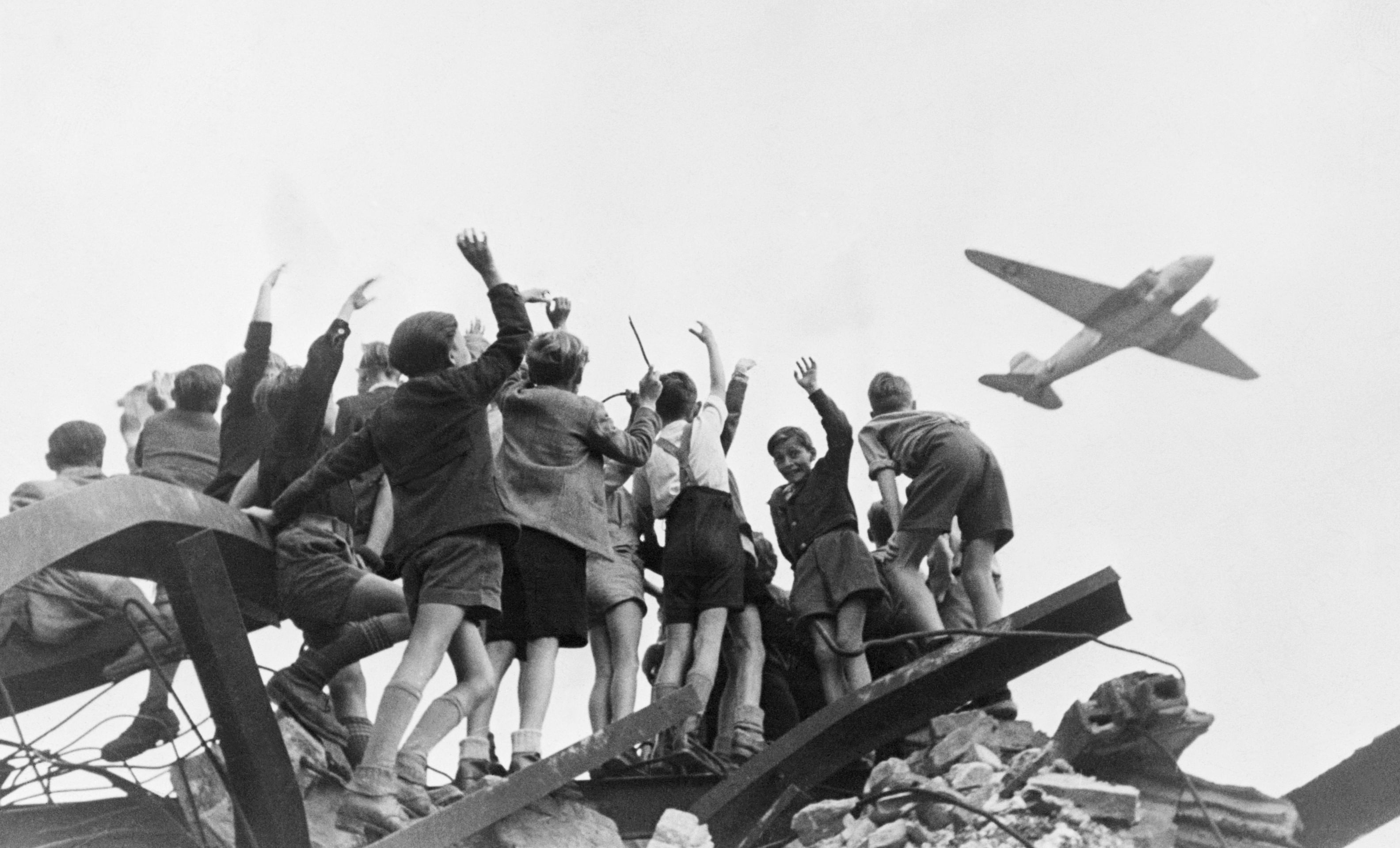 Es ist ein schwarz-weiß Foto. Eine Gruppe Kinder steht auf einem Berg von Schutt und Trümmern. Sie winken einem Flugzeug zu.