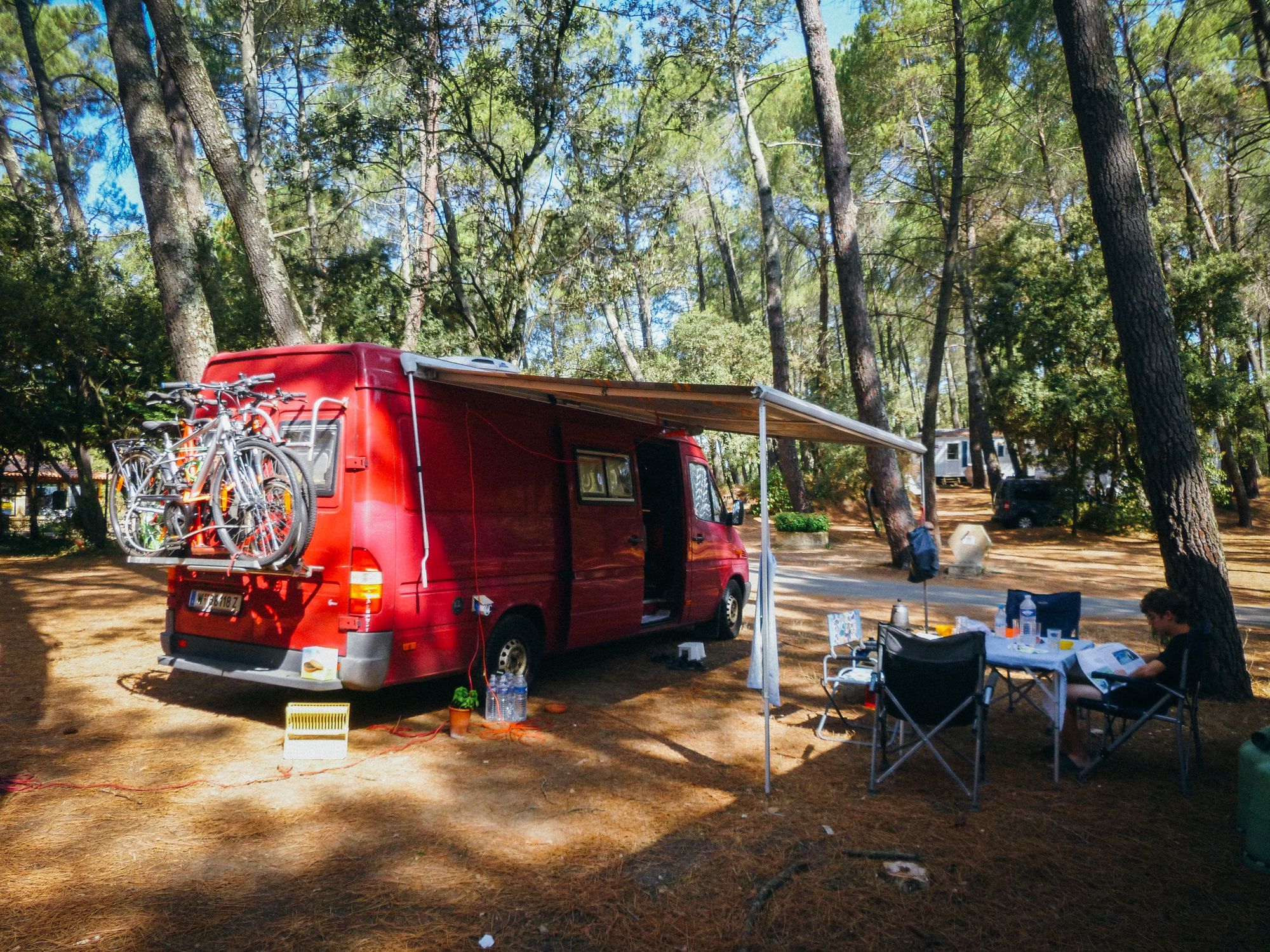 Ein roter Van steht inmitten von Bäumen. Davor ist ein Campingtisch aufgebaut. Ein Junge sitzt auf einem der Stühle.
