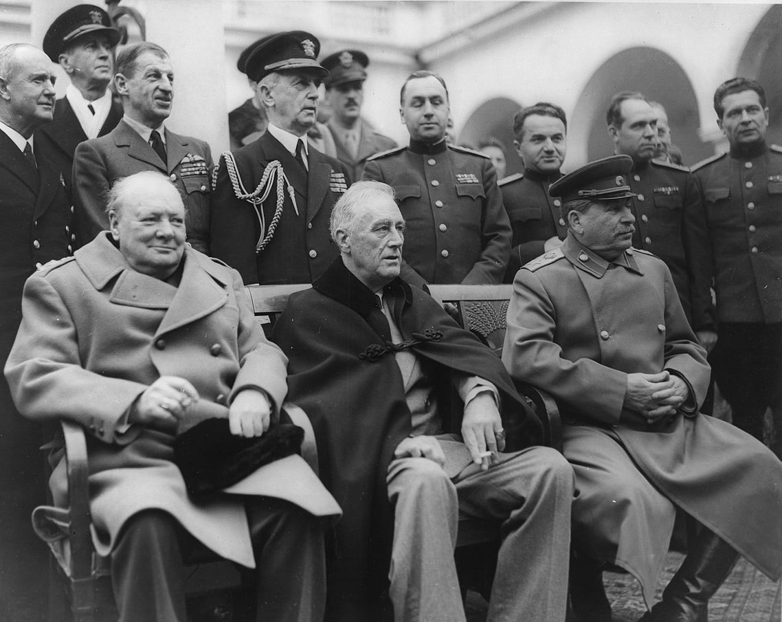 Churchill, Rosevelt und Stalin sitzen auf Stühlen nebeneinander. Im Hintergrund stehen hochrangige Militärs.