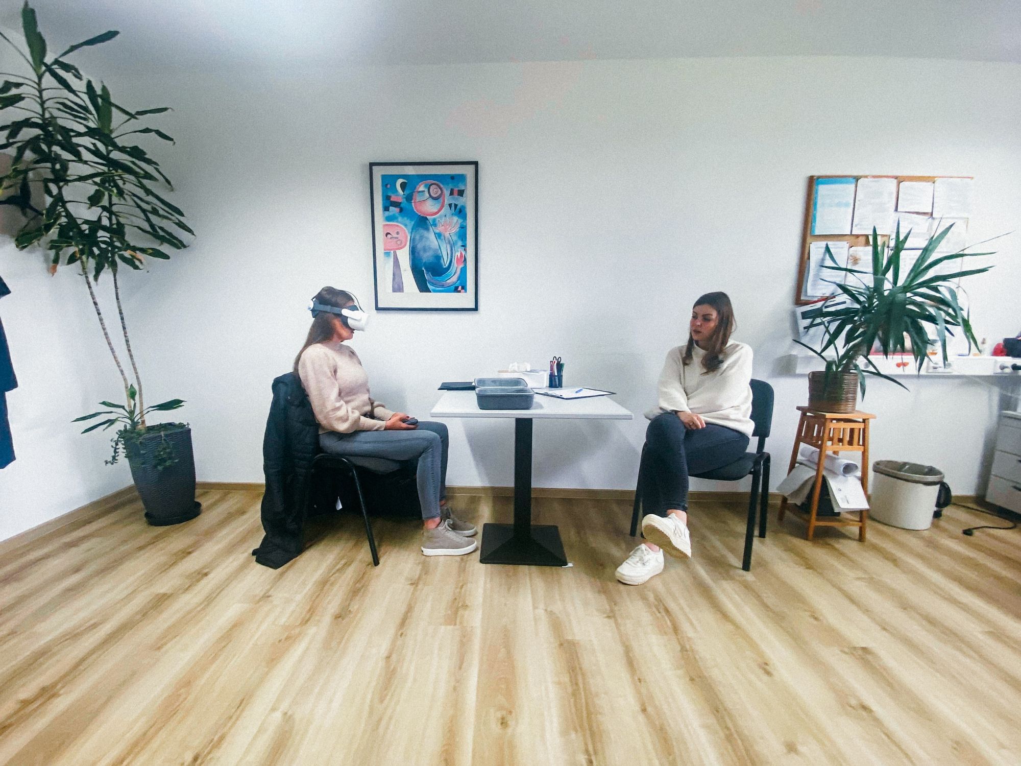 An einem Tisch sitzen sich eine Klinikangestellte und eine Patientin gegenüber. Die Patientin trägt eine VR-Brille.