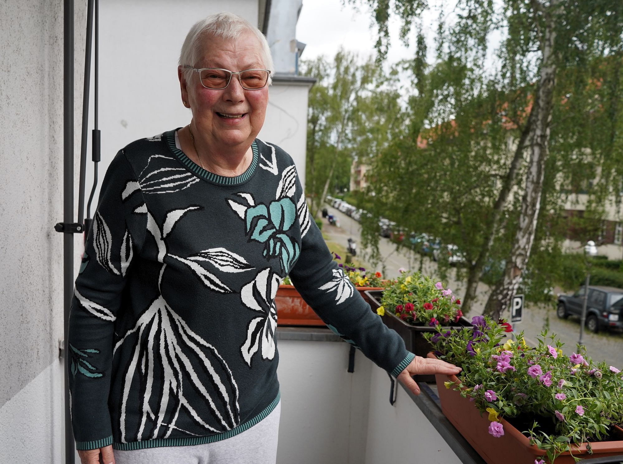 Birgit Krüger, eine ältere Dame mit kurzen grauen Haaren und Brille, steht auf einem Balkon und lächelt in die Kamera.