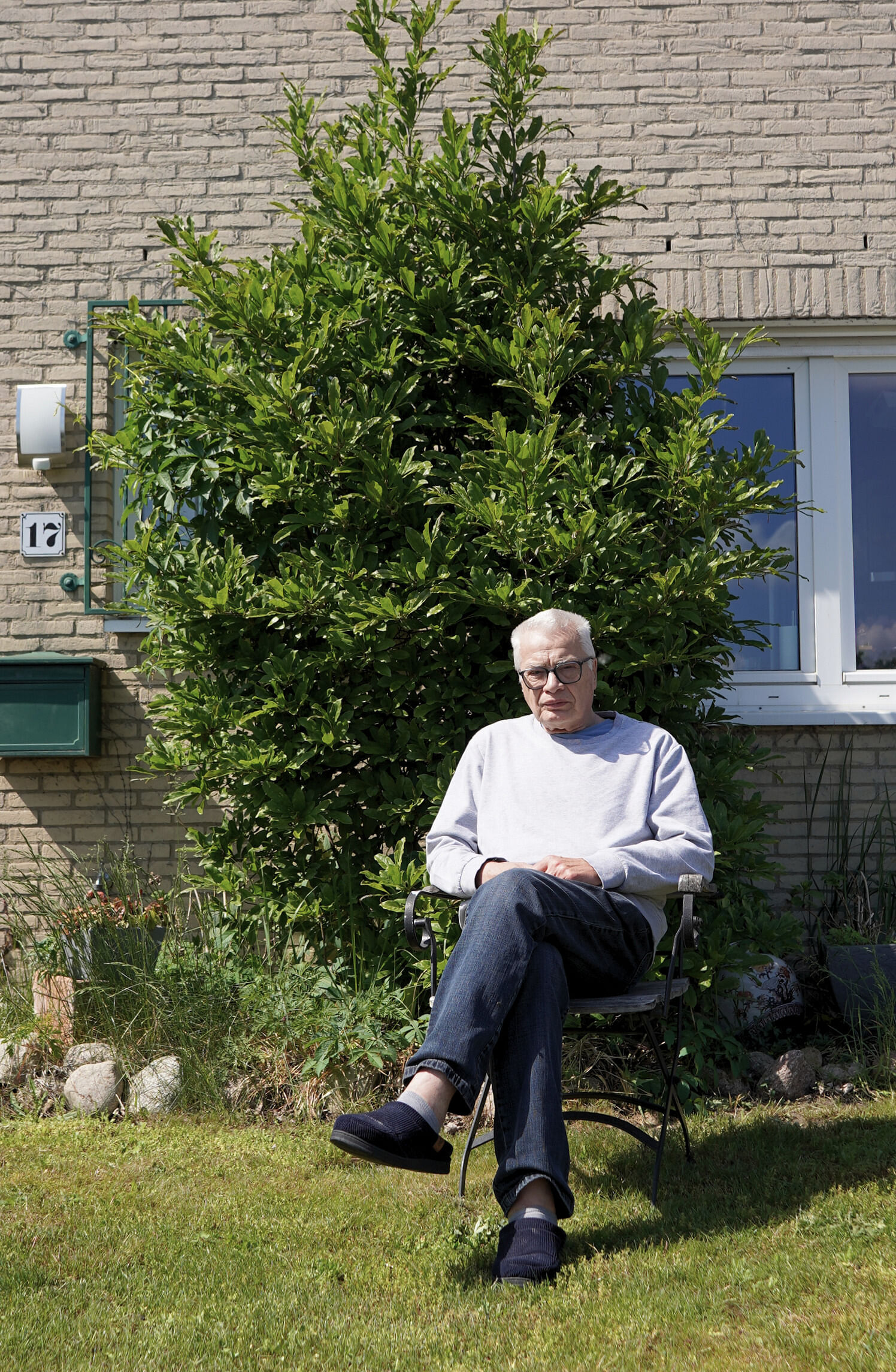 Erhard Neubert sitzt auf einem Stuhl vor seinem Haus. Hinter ihm ist ein Busch. Neubert trägt Jeans und einen grauen Pullover und eine Brille.