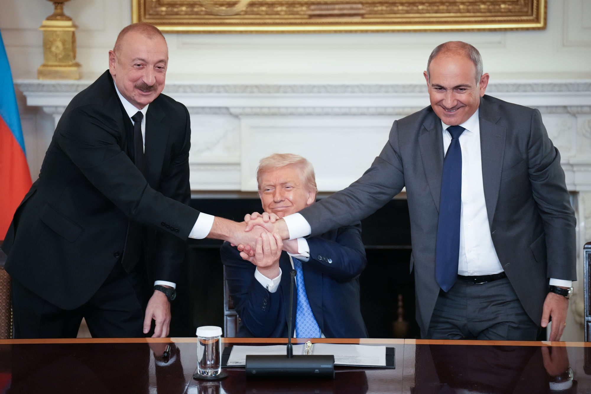 Auf dem Bild sind drei Männer zu sehen. In der Mitte sitzt Donald Trump, links steht Alijew, rechts steht Paschinjan. Sie halten alle gleichzeitig ihre Hände in der Mitte.