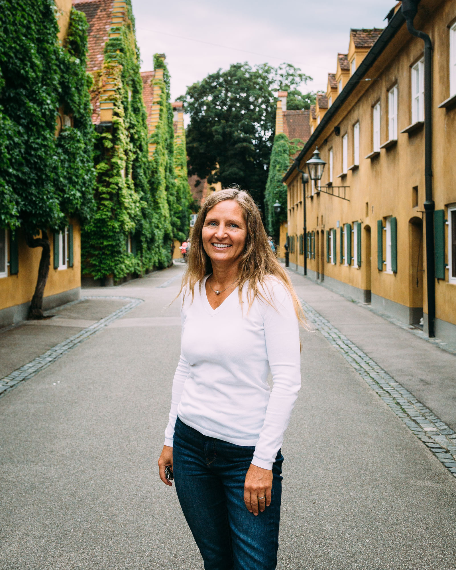 Eine Frau mit hellem Oberteil und Jeans steht auf einer Straße und lächelt in die Kamera.