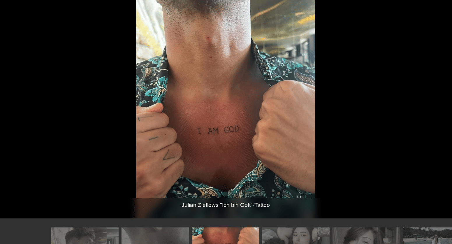 Das Bild zeigt Julian Zietlow, der sein Hemd zur Seite zieht, um ein Tattoo auf seiner Brust zu präsentieren. Das Tattoo besteht aus den Worten „I AM GOD“ in einfachen, schwarzen Großbuchstaben und befindet sich mittig unterhalb des Schlüsselbeins. Die Haut um das Tattoo wirkt leicht gerötet, was auf ein frisch gestochenes Tattoo hindeuten könnte. Am unteren Bildrand steht der Hinweis: „Julian Zietlows 'Ich bin Gott'-Tattoo“.