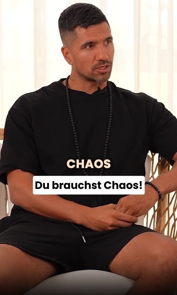 Auf dem Bild sitzt Julian Zietlow auf einem Sessel. Er trägt ein lockeres, schwarzes T-Shirt und eine kurze schwarze Hose. Um den Hals hängt eine lange schwarze Perlenkette. Seine Hände liegen locker übereinander auf seinem Schoß. Im Hintergrund sind helle, lichtdurchflutete Gardinen zu sehen. Über dem Bild steht der Schriftzug: „Du brauchst Chaos!“