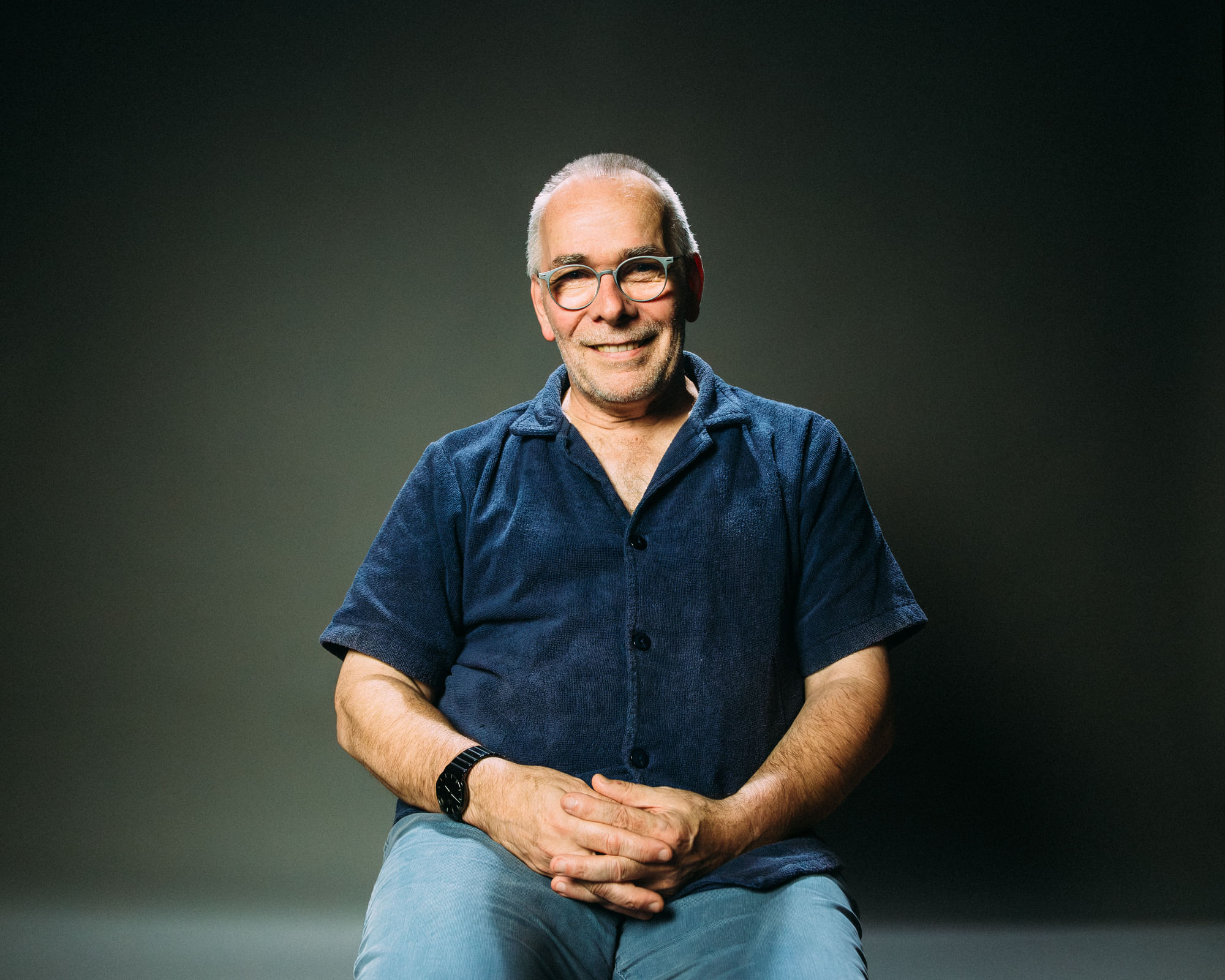 Ein Portraitbild von Martin Süsterhenn. Er sitzt auf einem Stuhl und schaut in die Kamera, der Hintergrund ist eine dunkelgrün-graue Fläche.