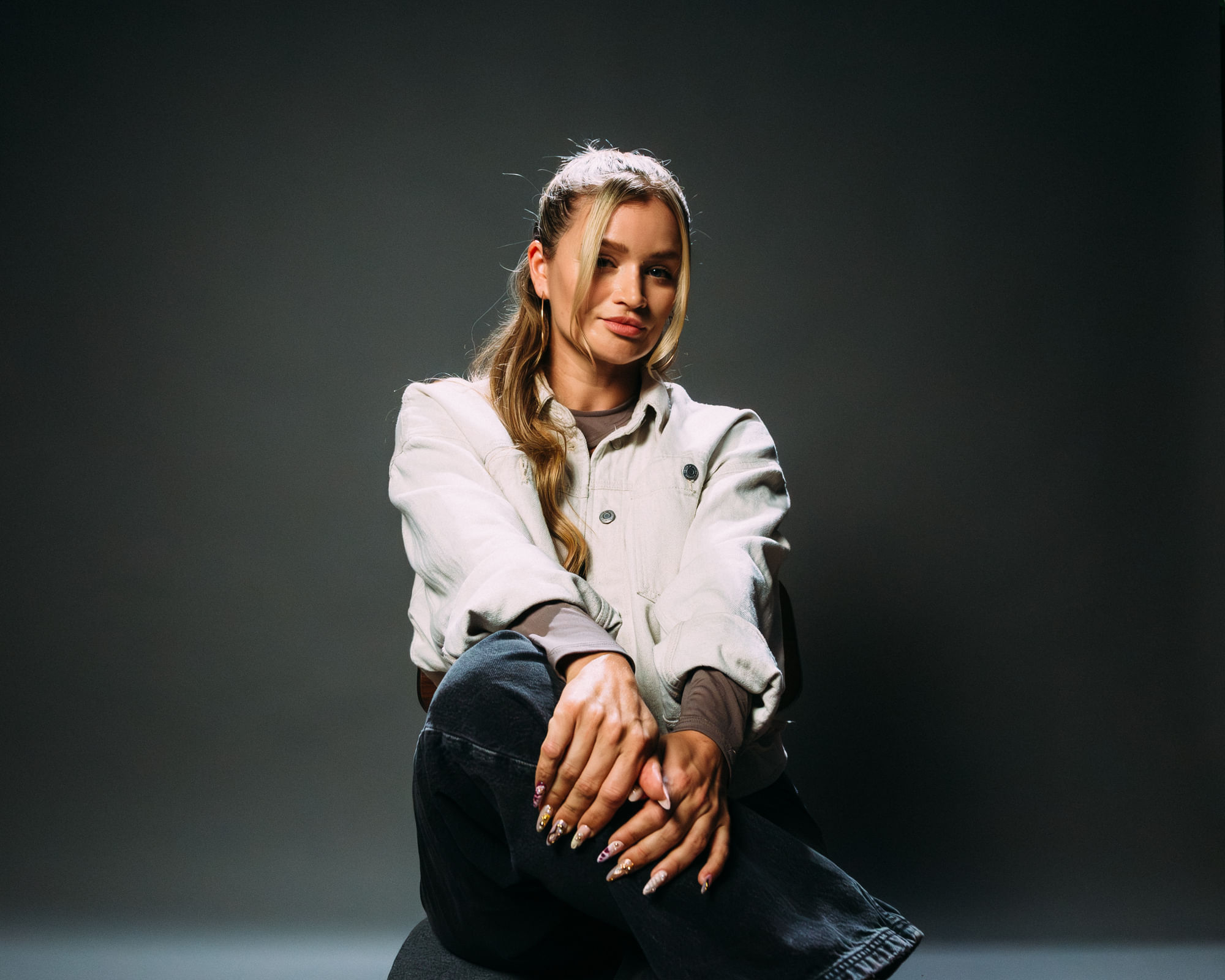 Portraitbild von Gina Waibel. Sie sitzt auf einem Stuhl, mit überschlagenen Beinen, und schaut in die Kamera. Der Hintergrund ist eine dunkelgrün-graue Fläche.