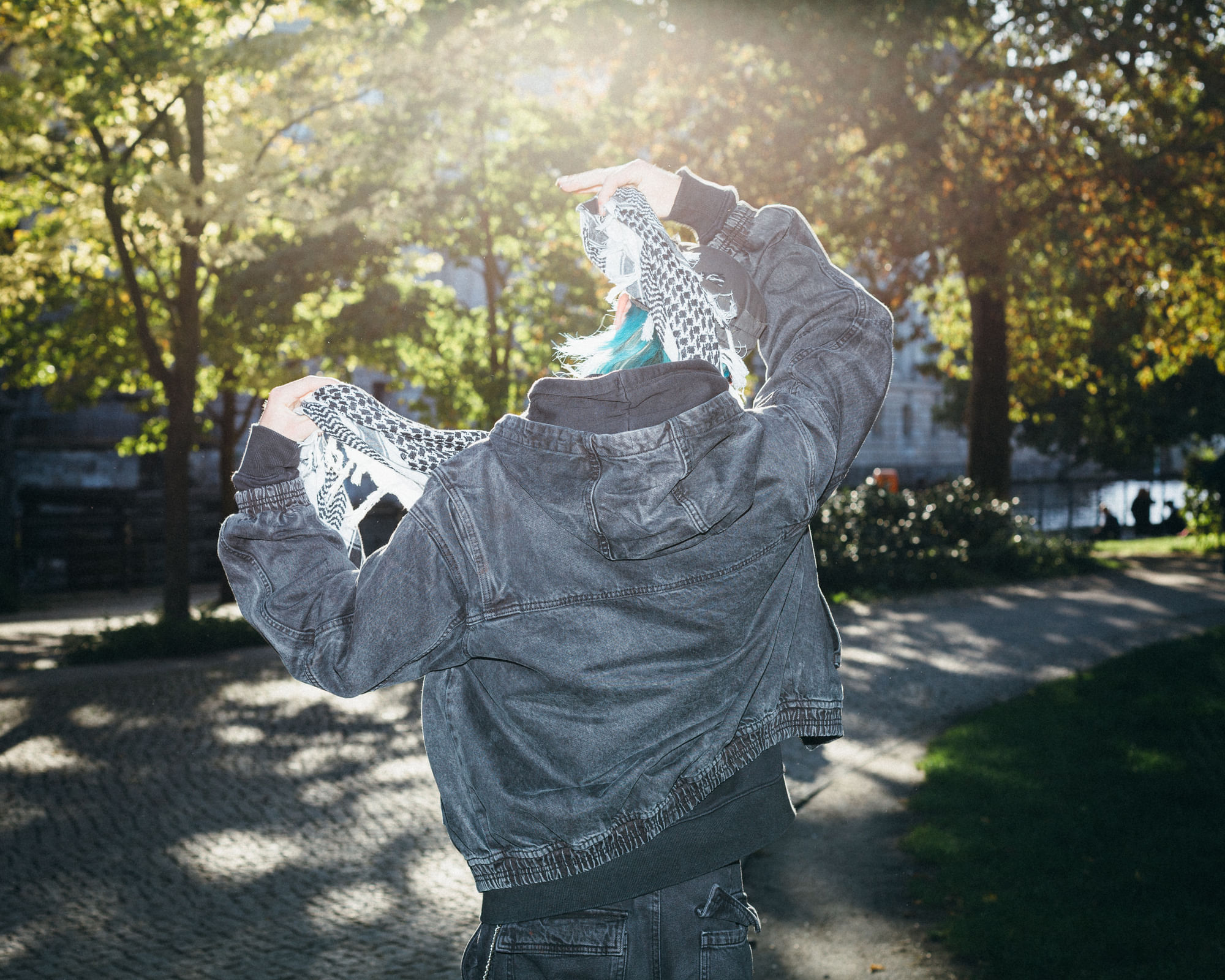 Ein Junge mit blauen Haaren von hinten, eine schwarze Jacke, in der Hand ein Palästinensertuch.