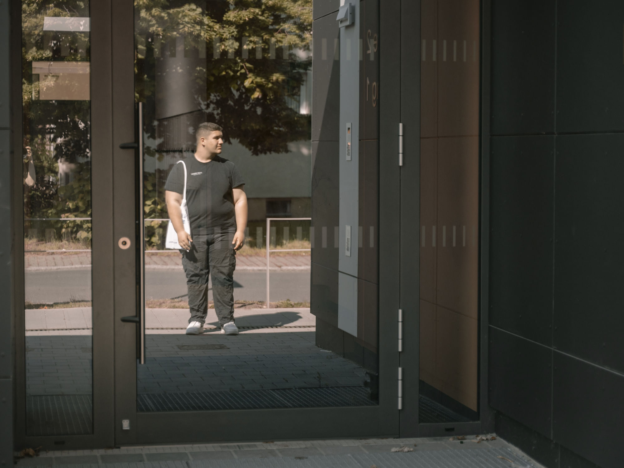 Ein Junge spiegelt sich in einer Tür, er trägt eine schwarze Hose und ein schwarzes T-Shirt und hat einen Leinenbeutel umhängen.