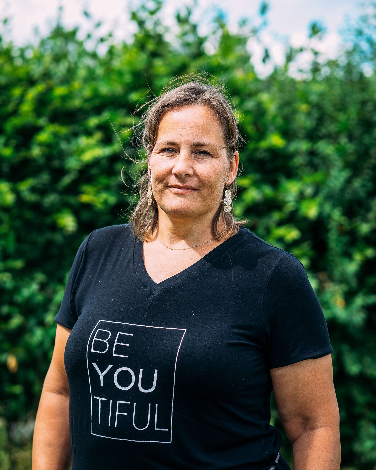 Eine Frau in einem schwarzen T-Shirt blickt in die Kamera. Sie steht vor einer Hecke.