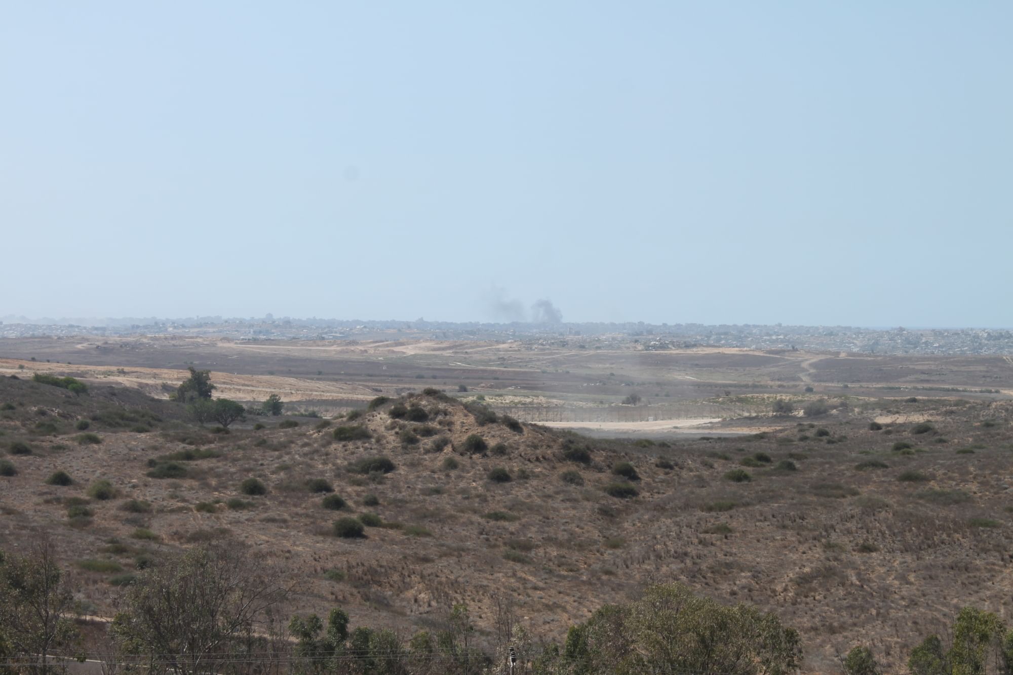 Man sieht eine karge Landschaft, im Hintergrund die Ruinen einer Stadt. Es steigen Rauchwolken auf.