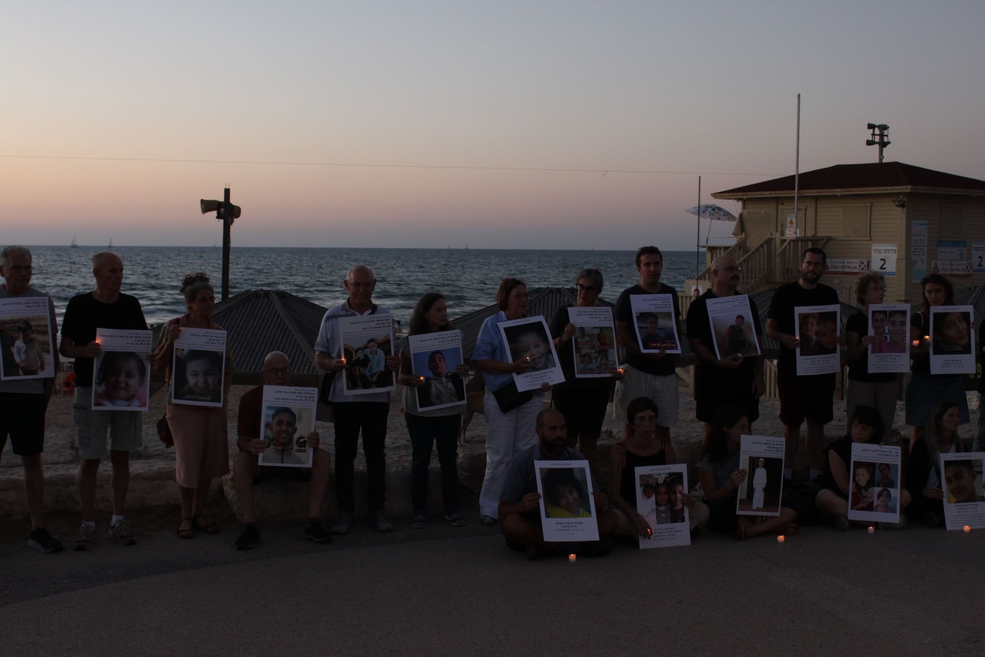Auf dem Foto sieht man eine Gruppe von Menschen, die im Sonnenuntergang an einer Promenade stehen und Fotos von Kindern in der Hand halten.