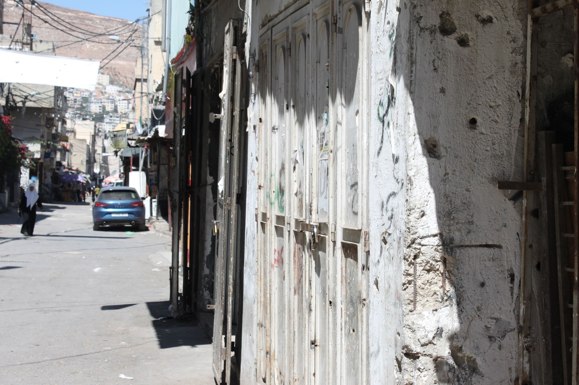 Man sieht eine Straße mit einem parkenden Auto und eine Hauswand mit Einschusslöchern.
