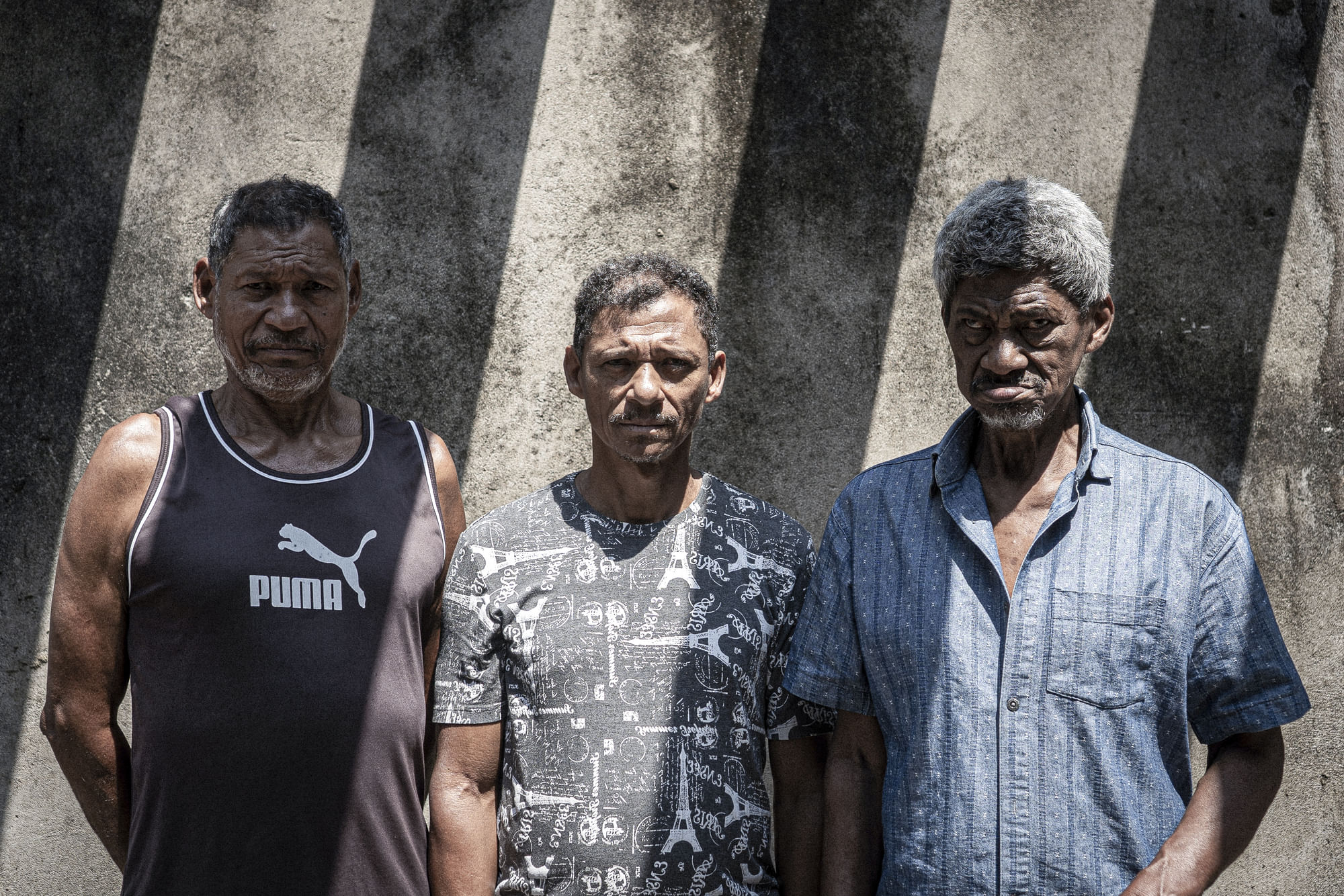 Drei Menschen stehen nebeneinander und blicken in die Kamera. Das Sonnenlicht wirft einen gestreiften Schatten über das gesamte Bild.